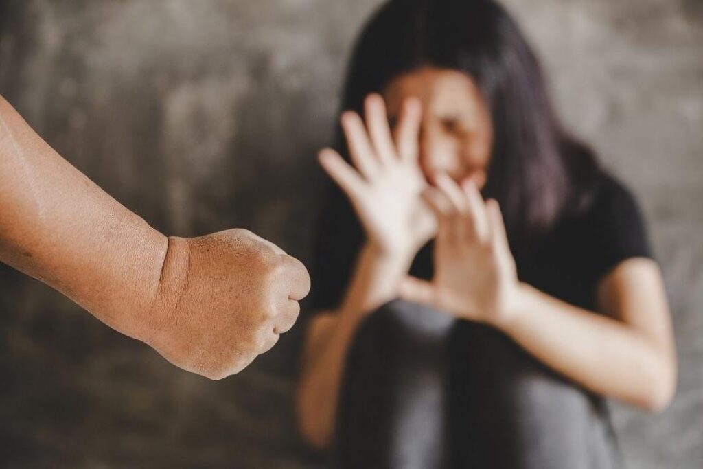 Donna rannicchiata e pugno di un uomo in primo piano, simbolo di maltrattamenti